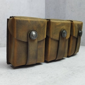 Puede incluir: Un conjunto de tres bolsas de cuero marrón con aspecto envejecido. Cada bolsa tiene una solapa asegurada por una correa y un botón metálico decorativo. Las bolsas están dispuestas una al lado de la otra.