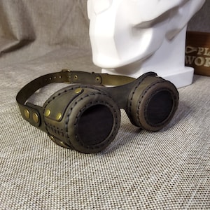 May include: Steampunk-style goggles with dark brown leather and brass accents. The goggles feature circular lenses with a mesh screen and are connected by a leather strap. The design includes decorative stitching and brass rivets, giving them a vintage, industrial look.
