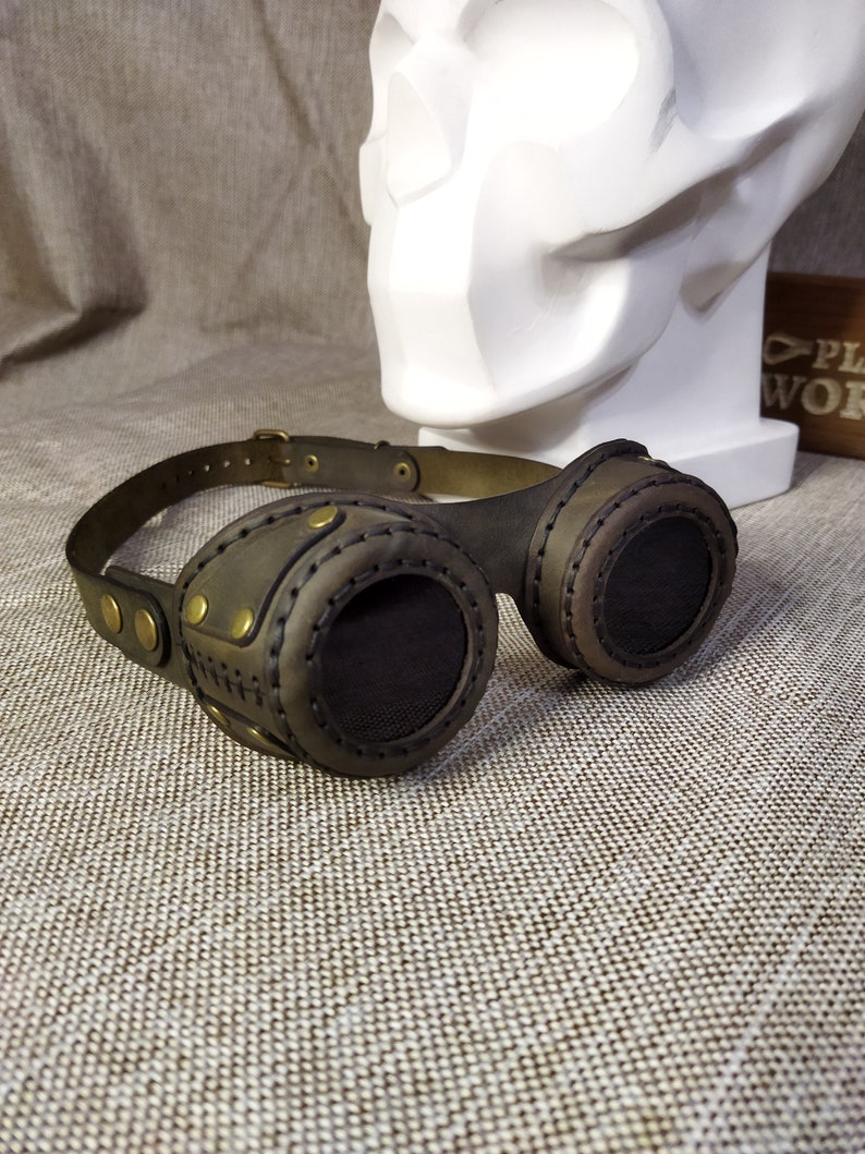 May include: Steampunk-style goggles with dark brown leather and brass accents. The goggles feature circular lenses with a mesh screen and are connected by a leather strap. The design includes decorative stitching and brass rivets, giving them a vintage, industrial look.
