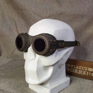 May include: Steampunk-style goggles with dark brown leather and olive green accents. The goggles feature circular lenses and are attached to a dark green adjustable strap. The goggles are displayed on a white bust.