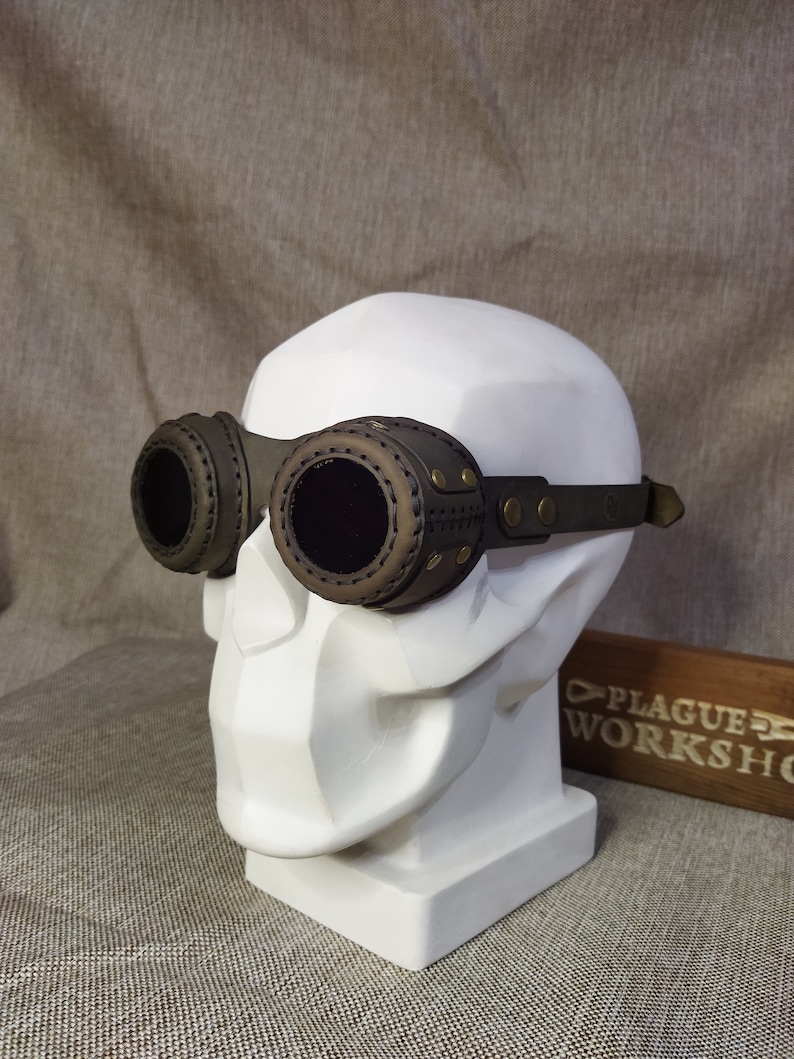 May include: Steampunk-style goggles with dark brown leather and olive green accents. The goggles feature circular lenses and are attached to a dark green adjustable strap. The goggles are displayed on a white bust.