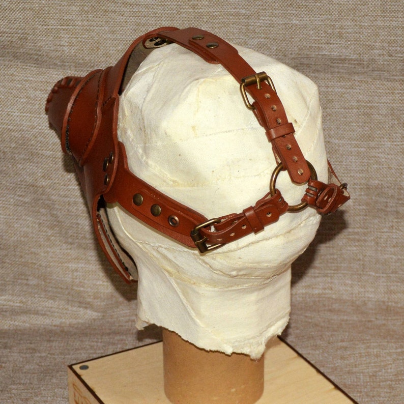 K&ouml;nnte beinhalten: Eine braune Ledermaske mit schnabelartigem Design, mit verstellbaren Riemen und Messingschnallen. Die Maske ist so konzipiert, dass sie &uuml;ber das Gesicht passt und mit Riemen um den Kopf befestigt wird. Die Maske wird auf einer wei&szlig;en, umwickelten Form pr&auml;sentiert.
