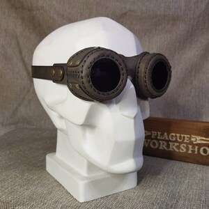 May include: Steampunk-style goggles with dark lenses and a brown leather frame. The goggles are mounted on a white bust. The frame features decorative stitching and brass-colored rivets. A wooden sign with the text "PLAGUE WORKSHOP" is in the background.