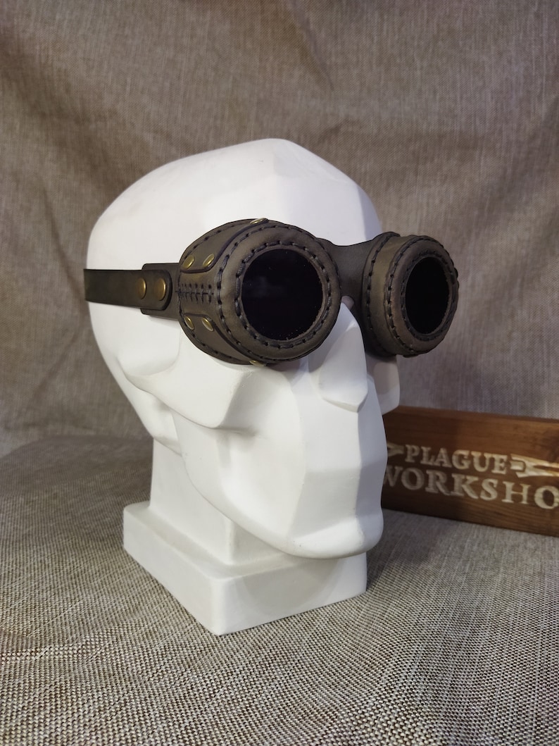 May include: Steampunk-style goggles with dark lenses and a brown leather frame. The goggles are mounted on a white bust. The frame features decorative stitching and brass-colored rivets. A wooden sign with the text "PLAGUE WORKSHOP" is in the background.