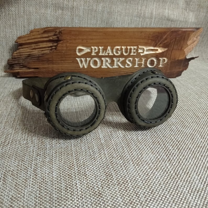 May include: Steampunk-style goggles with olive green leather and clear lenses. The goggles have a stitched design around the lenses and a dark leather strap. A wooden sign with the text "PLAGUE WORKSHOP" is in the background.