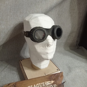 May include: Black steampunk goggles with circular lenses and a stitched leather frame. The goggles are displayed on a white head wrapped in bandages, mounted on a wooden base. The background is a gray fabric.
