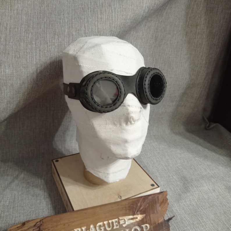 May include: Black steampunk goggles with circular lenses and a stitched leather frame. The goggles are displayed on a white head wrapped in bandages, mounted on a wooden base. The background is a gray fabric.
