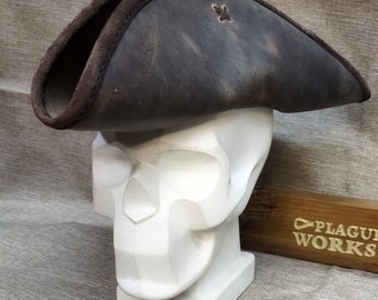May include: A dark brown leather tricorn hat with a brown suede brim, displayed on a white skull-shaped sculpture. The hat features a stitched cross detail. The wooden sign in the background reads "PLAGUE WORKS."