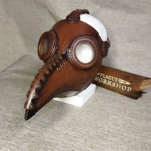 K&ouml;nnte beinhalten: Eine braune Leder-Pestdoktormaske mit langem Schnabel, kreisf&ouml;rmigen Augen&ouml;ffnungen und Metallakzenten. Die Maske hat eine wei&szlig;e Oberseite und wird auf einem wei&szlig;en Block pr&auml;sentiert. Der Text "PLAGUE WORKSHOP" ist auf einem Holzst&uuml;ck zu sehen.