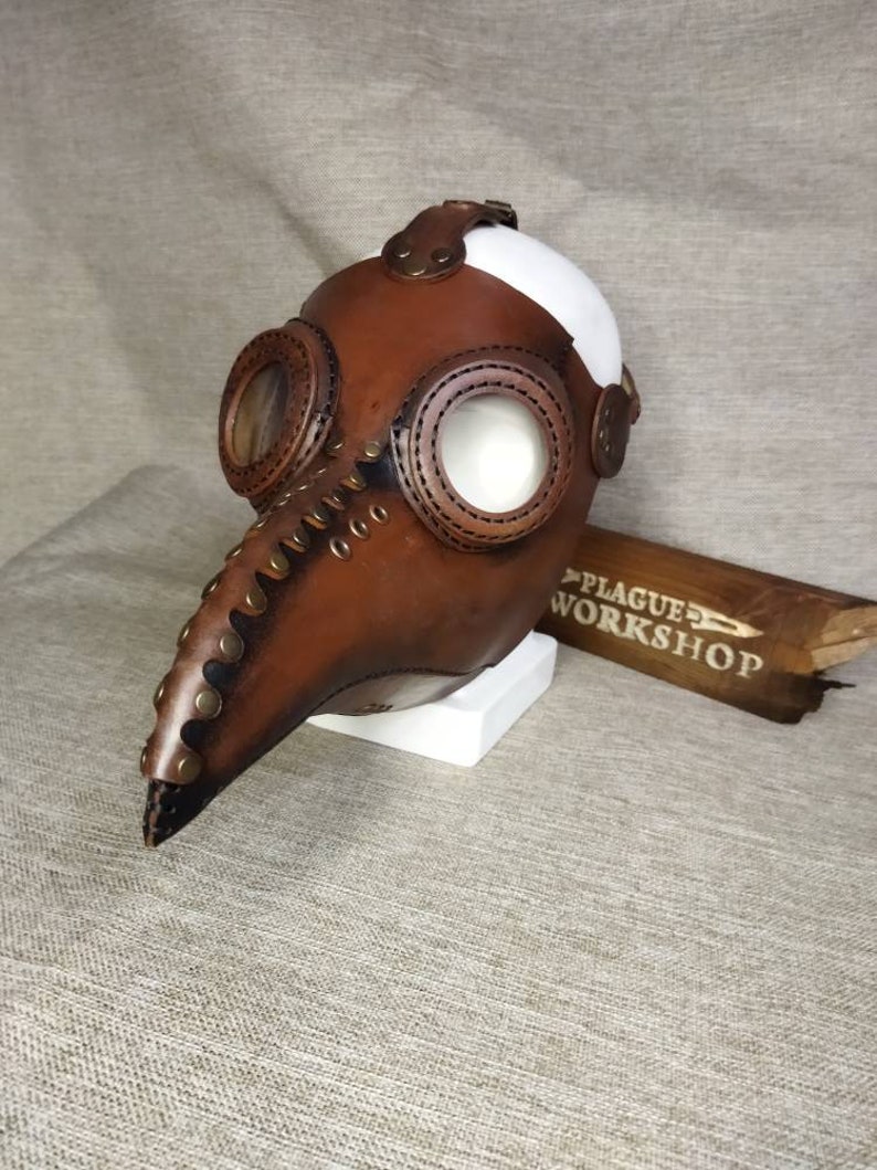 K&ouml;nnte beinhalten: Eine braune Leder-Pestdoktormaske mit langem Schnabel, kreisf&ouml;rmigen Augen&ouml;ffnungen und Metallakzenten. Die Maske hat eine wei&szlig;e Oberseite und wird auf einem wei&szlig;en Block pr&auml;sentiert. Der Text "PLAGUE WORKSHOP" ist auf einem Holzst&uuml;ck zu sehen.