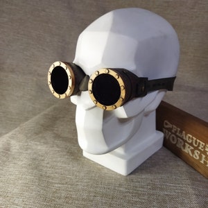 May include: Steampunk goggles featuring circular black lenses with gold-coloured rims. The goggles are attached to a dark brown leather strap and are displayed on a white skull-shaped bust. A wooden sign with "PLAGUE WORKSHOP" is visible.