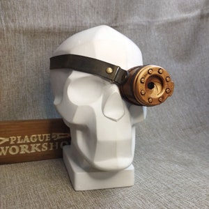 May include: Steampunk-style goggles with a bronze-colored lens and a dark brown leather strap. The goggles are attached to a white skull-shaped sculpture. The text "PLAGUE WORKSHOP" is visible on a wooden sign.