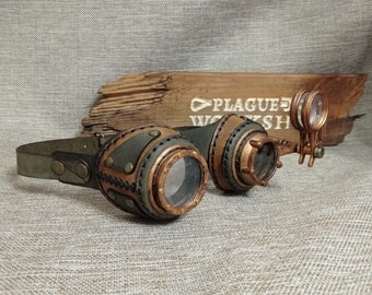May include: Steampunk goggles with a leather strap and frame. The goggles feature brown, green, and copper-coloured accents. The lenses are clear, and the design includes decorative stitching. The text "PLAGUE WORKSHOP" is visible on a wooden plank in the background.