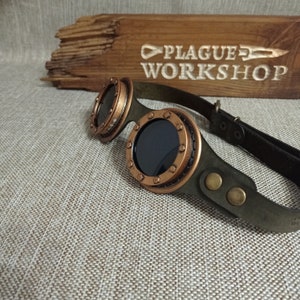 Könnte beinhalten: Steampunk-Brille mit dunklen Gläsern und kupferfarbenen Metallrahmen. Die Brille hat ein dunkelgrünes Lederband. Im Hintergrund befindet sich ein Holzschild mit der Aufschrift "PLAGUE WORKSHOP".