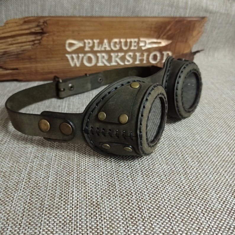 May include: Dark green leather steampunk goggles with brass rivets and stitching. The goggles have a wide adjustable strap and circular lenses. The text "PLAGUE WORKSHOP" is visible on a wooden sign in the background.