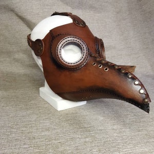 K&ouml;nnte beinhalten: Eine braune Leder-Pestdoktormaske mit klaren, kreisf&ouml;rmigen Augenlinsen. Die Maske hat einen langen, gebogenen Schnabel und ist mit Riemen und Nieten befestigt. Die Maske wird auf einer wei&szlig;en Kopfattrappe ausgestellt.
