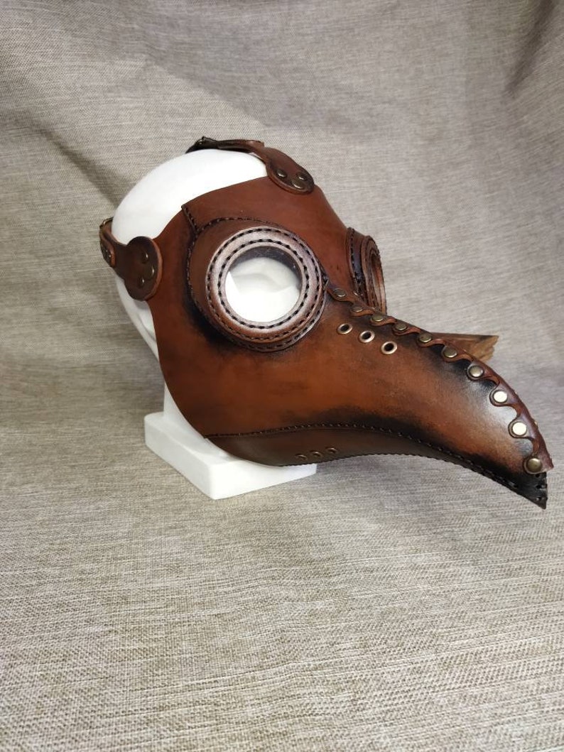 K&ouml;nnte beinhalten: Eine braune Leder-Pestdoktormaske mit klaren, kreisf&ouml;rmigen Augenlinsen. Die Maske hat einen langen, gebogenen Schnabel und ist mit Riemen und Nieten befestigt. Die Maske wird auf einer wei&szlig;en Kopfattrappe ausgestellt.