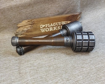 May include: Two replica grenades with wooden handles and metallic grey tops. The grenades are lying on a textured surface, with a wooden sign that reads "PLAGUE WORKSHOP" in the background.