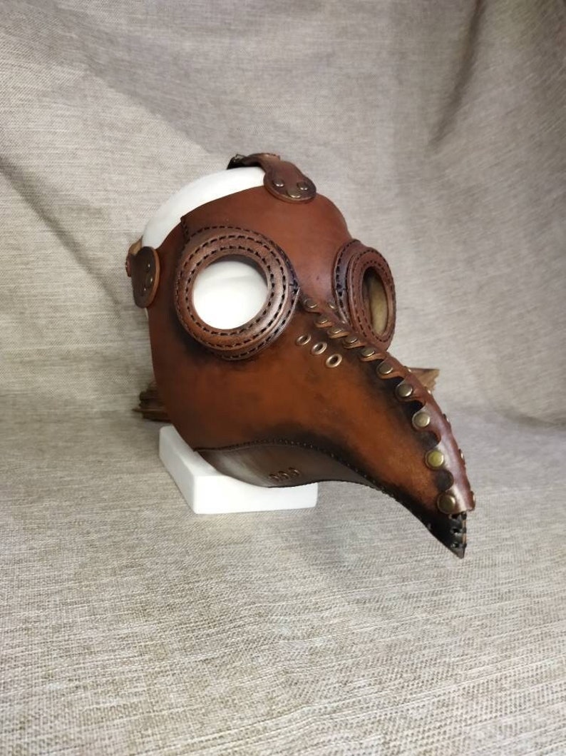 K&ouml;nnte beinhalten: Eine braune Leder-Pestdoktormaske mit langem Schnabel und kreisf&ouml;rmigen Augen&ouml;ffnungen. Die Maske hat Messingnieten und Nahtdetails, mit wei&szlig;em Innenraum und einem Riemen. Die Maske wird auf einem wei&szlig;en St&auml;nder pr&auml;sentiert.