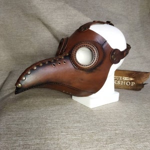 K&ouml;nnte beinhalten: Eine braune Leder-Pestdoktormaske mit langem, gebogenem Schnabel und gro&szlig;en, kreisf&ouml;rmigen Augen&ouml;ffnungen. Die Maske hat Nieten und Nahtdetails und wird auf einem wei&szlig;en Schaufensterkopf ausgestellt.