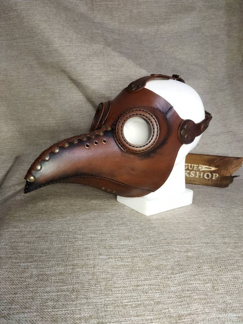 K&ouml;nnte beinhalten: Eine braune Leder-Pestdoktormaske mit langem, gebogenem Schnabel und gro&szlig;en, kreisf&ouml;rmigen Augen&ouml;ffnungen. Die Maske hat Nieten und Nahtdetails und wird auf einem wei&szlig;en Schaufensterkopf ausgestellt.