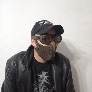 May include: A brown leather mask with a steampunk design, featuring circular eye goggles and a perforated front. The mask is worn with a black cap that says "PLAGUE WORKS" and a black leather jacket. The mask has a vintage, industrial aesthetic.