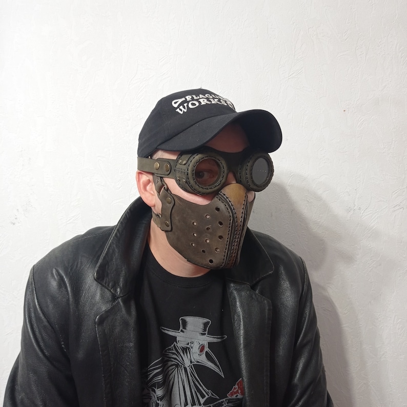 May include: A brown leather mask with a steampunk design, featuring circular eye goggles and a perforated front. The mask is worn with a black cap that says "PLAGUE WORKS" and a black leather jacket. The mask has a vintage, industrial aesthetic.