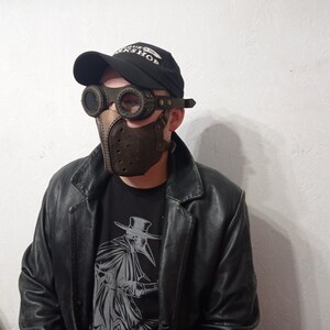 May include: A brown leather plague doctor mask with matching goggles and a black baseball cap. The mask has a beak-like design with small holes. The goggles have round lenses and are attached to the mask. The person is wearing a black leather jacket and a black t-shirt with a graphic.