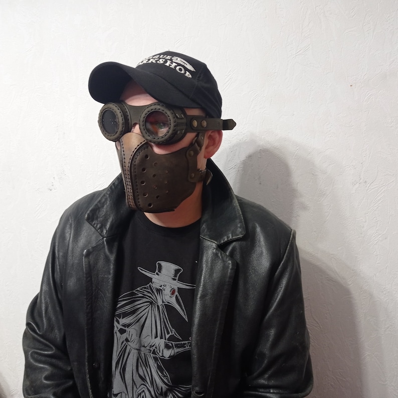 May include: A brown leather plague doctor mask with matching goggles and a black baseball cap. The mask has a beak-like design with small holes. The goggles have round lenses and are attached to the mask. The person is wearing a black leather jacket and a black t-shirt with a graphic.