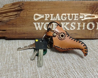 May include: A brown leather keychain shaped like a plague doctor mask, with black stitching and metal hardware. The keychain is attached to a keyring with several keys. The background features a wooden sign with the words "PLAGUE WORKSHOP" carved into it.