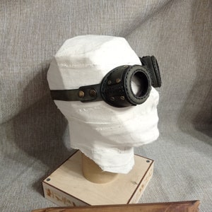May include: Steampunk-style goggles with dark green leather and brass accents. The goggles feature round lenses and are attached to a head strap. The design includes decorative stitching and rivets, giving them a vintage, industrial look.