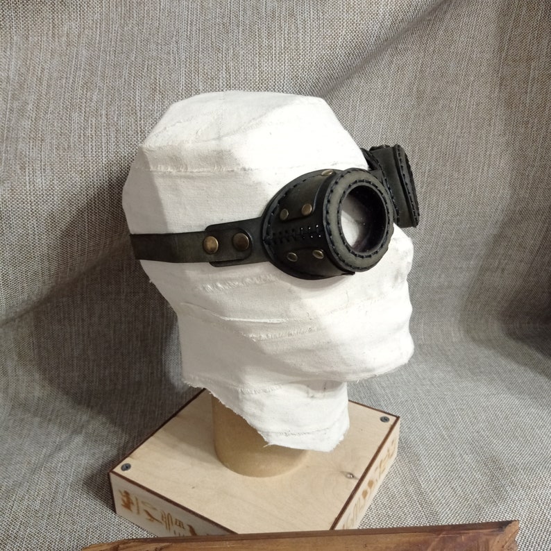 May include: Steampunk-style goggles with dark green leather and brass accents. The goggles feature round lenses and are attached to a head strap. The design includes decorative stitching and rivets, giving them a vintage, industrial look.