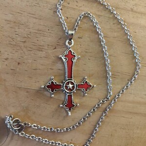 Pode incluir: Um colar de corrente de prata com um pingente de cruz vermelha e prateada. A cruz tem uma estrela de cinco pontas no centro.