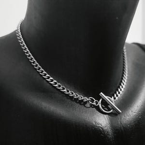 May include: A silver curb chain necklace with a toggle clasp. The necklace is displayed on a black mannequin, highlighting the chain links and the clasp design. A simple, stylish accessory. The chain is made of metal.
