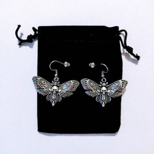 May include: A pair of silver moth earrings with a skull design, displayed on a black velvet pouch.