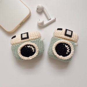 May include: Two crochet camera-shaped AirPod cases in light green, cream, black, and white. The cases have a lens design and a square viewfinder. White wireless earbuds and a smartphone are also in the image.