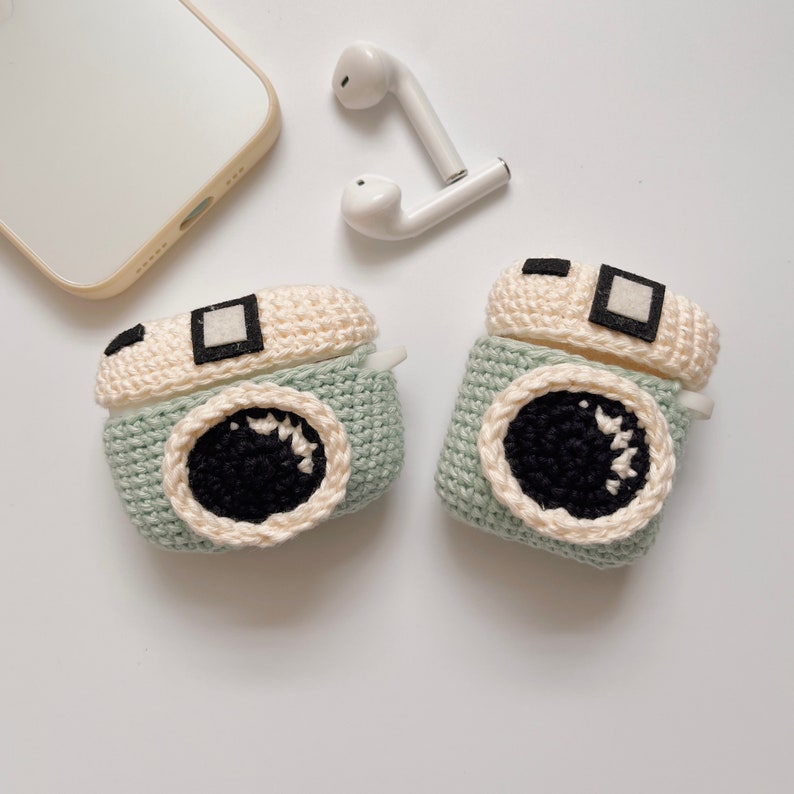 May include: Two crochet camera-shaped AirPod cases in light green, cream, black, and white. The cases have a lens design and a square viewfinder. White wireless earbuds and a smartphone are also in the image.