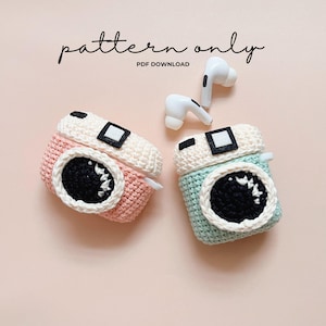 May include: Crocheted AirPod cases in the shape of cameras, one pink and one teal, with black and white details. The cases have a circular lens design and a square viewfinder. The text "pattern only PDF download" is visible.