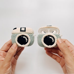 May include: Crocheted camera-shaped AirPod cases in light green, cream, and black. One case is closed, the other open, revealing white earbuds. The cases feature a circular lens design and a rectangular top detail, resembling a vintage camera.