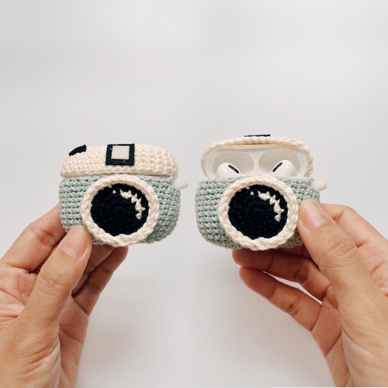 May include: Crocheted camera-shaped AirPod cases in light green, cream, and black. One case is closed, the other open, revealing white earbuds. The cases feature a circular lens design and a rectangular top detail, resembling a vintage camera.