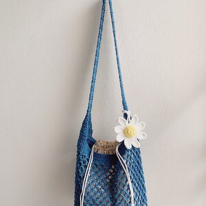 Peut inclure: Un sac cabas en crochet bleu avec une fleur de marguerite blanche en crochet attach&eacute;e sur le c&ocirc;t&eacute;. Le sac a une fermeture &agrave; cordon et une longue sangle.