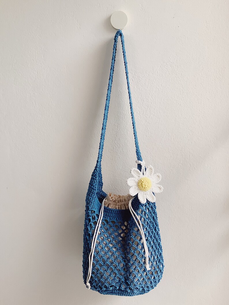 Peut inclure: Un sac cabas en crochet bleu avec une fleur de marguerite blanche en crochet attach&eacute;e sur le c&ocirc;t&eacute;. Le sac a une fermeture &agrave; cordon et une longue sangle.