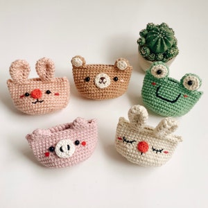 Puede incluir: Cinco bolsas de animales de crochet con caras bordadas. Las bolsas son marrones, rosas, verdes, blancas y beige. Los animales son un oso, un cerdo, una rana, un conejo y otro conejo.