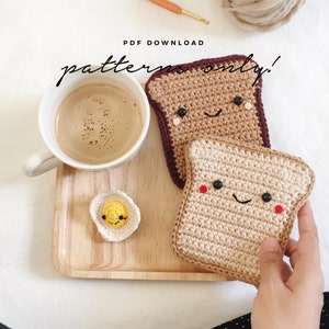 Pode incluir: Padrão de crochê para um conjunto de duas fatias de pão sorridentes e um ovo frito. O padrão está disponível para download em PDF. As fatias de pão são marrons e bege com um rubor vermelho nas bochechas. O ovo é amarelo com uma gema branca.