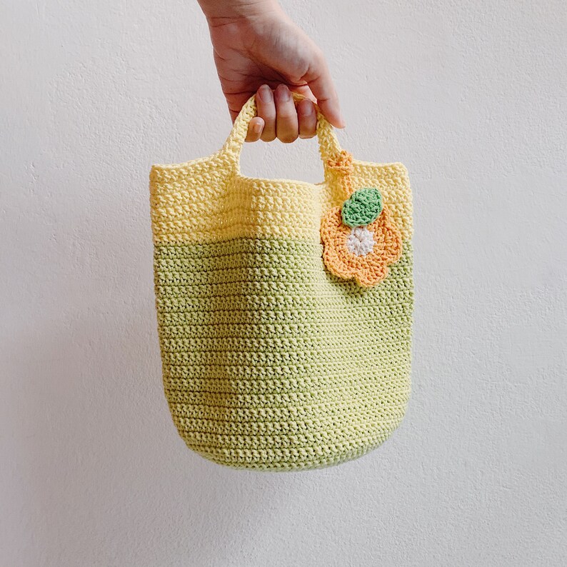 Peut inclure: Un sac fourre-tout en crochet avec un d&eacute;grad&eacute; de couleurs jaune et vert. Le sac est orn&eacute; d'une fleur en crochet avec des accents orange, blanc et vert.