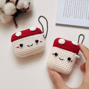 May include: Two crocheted mushroom-shaped AirPod cases with white bodies and red tops. Each case has a smiling face with black eyes and a black mouth.