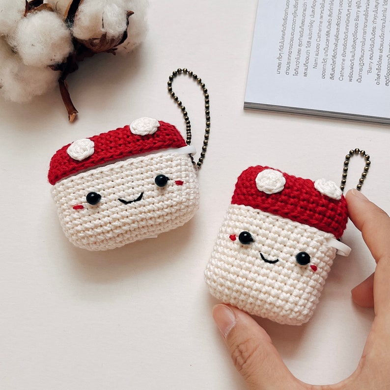 May include: Two crocheted mushroom-shaped AirPod cases with white bodies and red tops. Each case has a smiling face with black eyes and a black mouth.