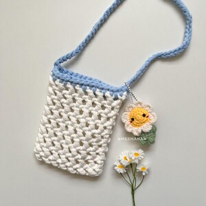 Pu&ograve; includere: Una borsa a tracolla all'uncinetto bianca con cinturino e finiture azzurre. Un piccolo ciondolo a forma di fiore all'uncinetto sorridente pende dalla tracolla. La borsa &egrave; a trama aperta e il testo "@MEEMANAN" &egrave; visibile.