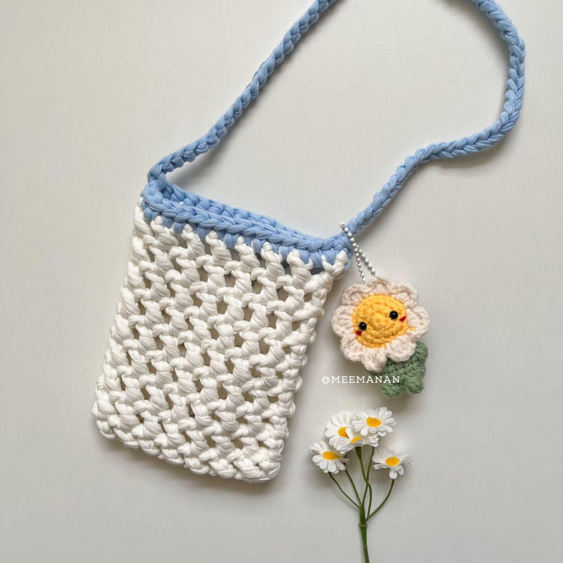 Pu&ograve; includere: Una borsa a tracolla all'uncinetto bianca con cinturino e finiture azzurre. Un piccolo ciondolo a forma di fiore all'uncinetto sorridente pende dalla tracolla. La borsa &egrave; a trama aperta e il testo "@MEEMANAN" &egrave; visibile.