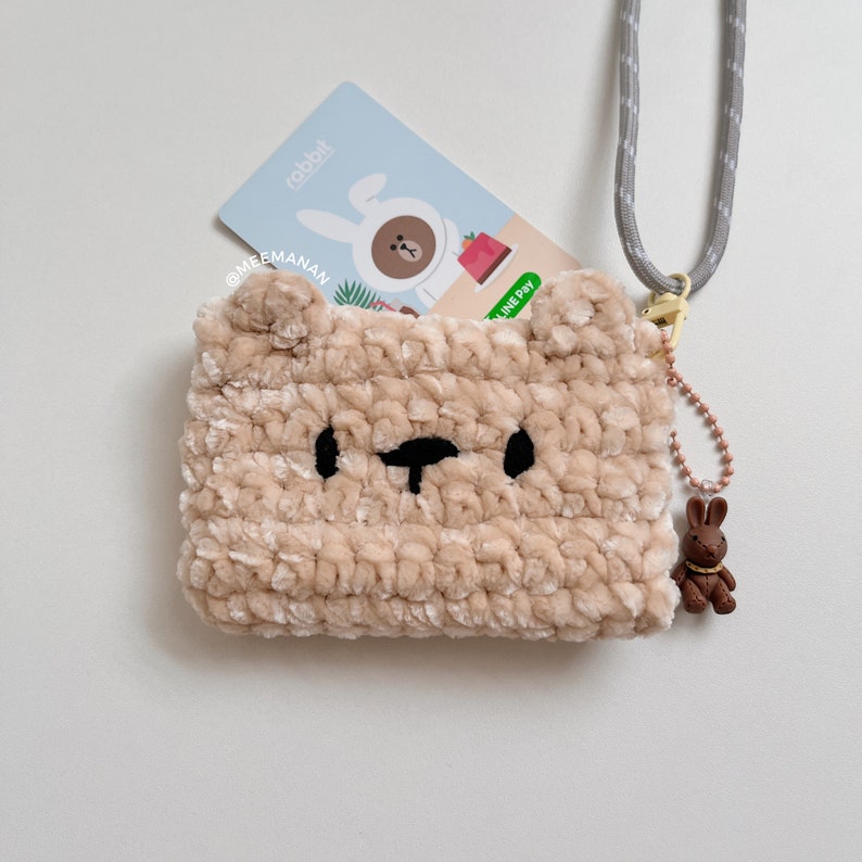 K&ouml;nnte beinhalten: Beige, geh&auml;kelte B&auml;ren-f&ouml;rmige Tasche mit schwarzen Augen und Nase. Eine Karte mit Kaninchen-Grafik ist teilweise sichtbar. Ein grauer Kordelriemen und ein brauner Hasen-Anh&auml;nger sind befestigt.